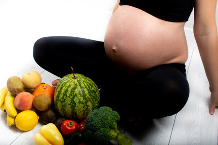 Pregnant woman with fruit and vegetables around her. Healthy eating concept. Pregnancy, slim skinny girl in black top and leggings isolated on white background. Diet and freshnessの写真素材
