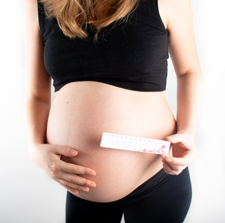 Young pregnant woman measures her waist with measurement tape, weight diet and healthy lifestyle pregnancy concept. Black top and leggings isolated model on white backgroundの写真素材