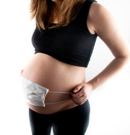 Pregnant woman holding white medicinal face mask on her pregnant belly abdomen. Coronavirus covid19 new normal rules and regulations, isolation and new lifestyle conceptの写真素材