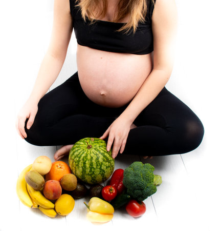 Pregnant woman with fruit and vegetables around her. Healthy eating concept. Pregnancy, slim skinny girl in black top and leggings isolated on white background. Diet and freshnessの写真素材