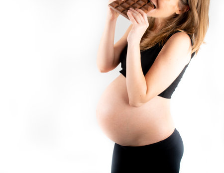 Pregnant woman holding a big dark chocolate bar. Gestational diabetes concept. Sugar free diet, unhealthy, healthy lifestyle. Slim fit skinny woman posing on white background. Black top and leggingsの写真素材