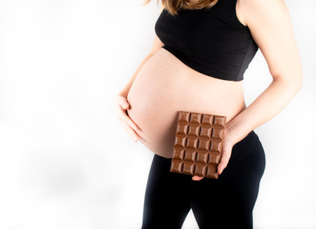 Pregnant woman holding a big dark chocolate bar. Gestational diabetes concept. Sugar free diet, unhealthy, healthy lifestyle. Slim fit skinny woman posing on white background. Black top and leggingsの写真素材