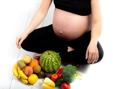 Pregnant woman with fruit and vegetables around her. Healthy eating concept. Pregnancy, slim skinny girl in black top and leggings isolated on white background. Diet and freshnessの写真素材