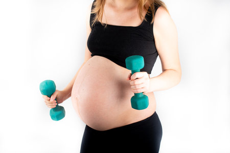 Active healthy pregnant woman exercising with dumbells, isolated on white background. The young expecting mother holding baby in pregnant belly. Maternity prenatal care and woman pregnancy concept.の写真素材