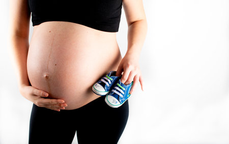 Pregnant woman holding baby boy small shoes in hands, happy pregnancy expecting gift concept. Closeup of mother to be isolated on white backgroundの写真素材