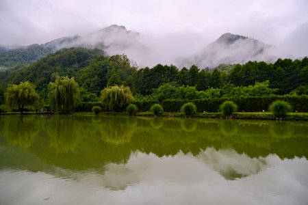 Fog and mist covering mountain peaks with reflection in calm green lake.の写真素材