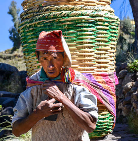 TAQUILE ISLAND, PUNO, PERU. MAY 31, 2013: Unidentified single man in traditional clothes carrying big rattan baskets, on the Taquile island, in Titicaca lakeのeditorial素材