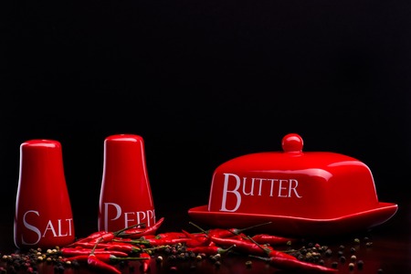 Closup of red gloss, ceramic salt-cellar, pepper-box and butter dish with chili and black pepper standing on dark wood table on a black backgroundの写真素材