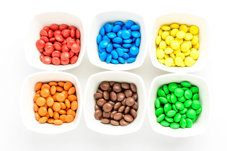 Six white squared bowls with small red, yellow, green, blue, brown and orange coated chocolate...