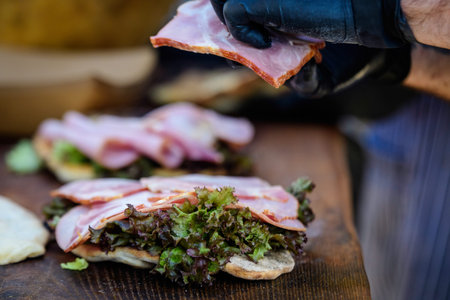 Hands of a man preparing a fresh healthy sandwich or burger with ham, green salad on a brown wooden...