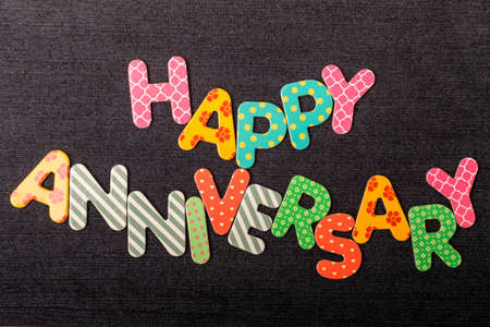 Card with Happy Anniversary words made from mixed vivid colored wooden letters on a textured dark black textile material that can be used as a messageの写真素材