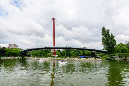 Bucharest, Romania - 29 May 2021: Landscape with the modern metallic bridge, lake and green trees in Drumul Taberei Park (Parcul Drumul Taberei) also known as Moghioros Park, in a cloudy summer dayのeditorial素材