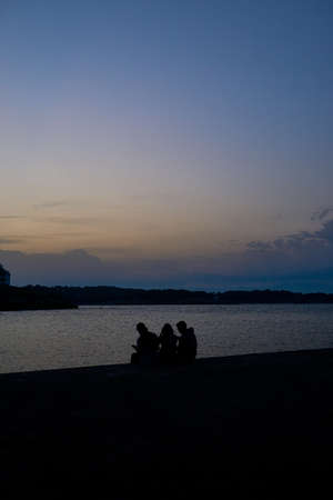 Silhouettes of three people sitting near the sea against a sunsetの写真素材