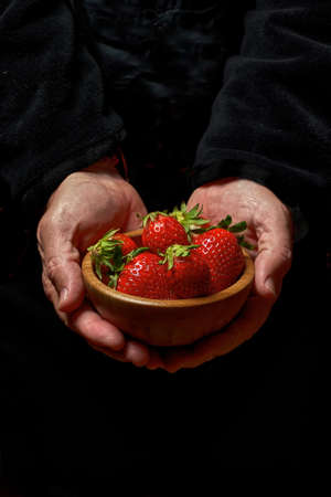 Man's hands holding a wooden bowl full of fresh strawberries on a black colored backgroundの写真素材