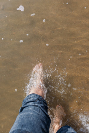 A person's foot in the water and the sand on the beach is brown. The person is standing on the beach, and the water is calm. The beach is sandyの写真素材