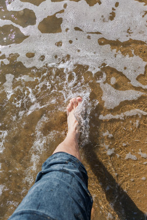 A person's foot in the water and the sand on the beach is brown. The person is standing on the beach, and the water is calm. The beach is sandyの写真素材
