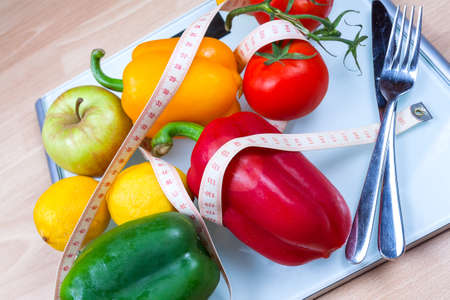 Peppers, tomatoes, apple and lemons on a scale with cutlery and tape measure. Abstract of healthy eating life.の写真素材