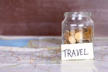 Coins in a jar with travel sign on a map against vintage brown wooden background. Copy space on the left. Concept of travelling.の写真素材