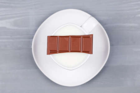 Bar of milk chocolate on a white cup of milk on grey natural wood background. Top view. Concept of making hot drink for autumn fall season.の写真素材