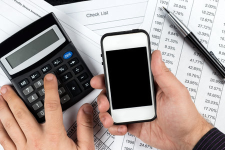 Hands of businessman holding white smartphone and calculating revenue or expenses on black...