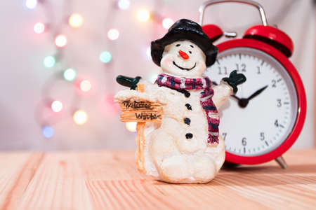 Little snowman figurine with red vintage clock. Natural wooden table. Christmas tree lights bokeh background. Copy space to the left.の写真素材
