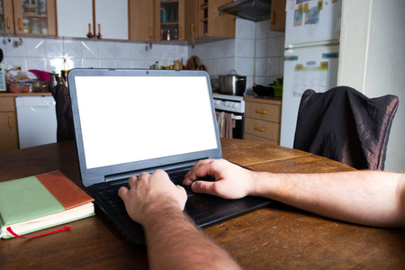 Man typing on a laptop computer in vintage kitchen. Blank white screen ready for mockup. Notepad....