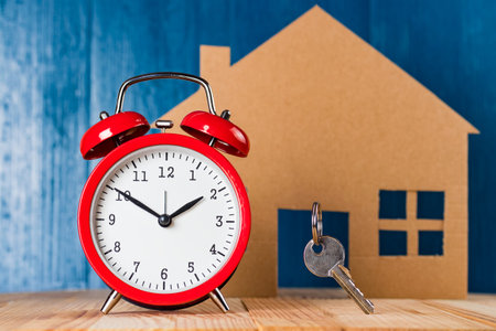 Old vintage alarm clock and door key against house shape and blue background. Time to buy a home...