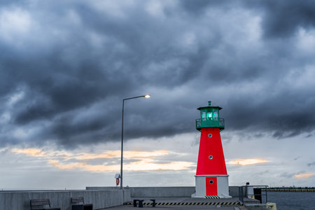 Red and green lighthouse on a harbor pier against dramatic clouds in the sky.
