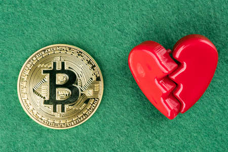 Gold physical Bitcoin coin with broken heart on green background.の写真素材