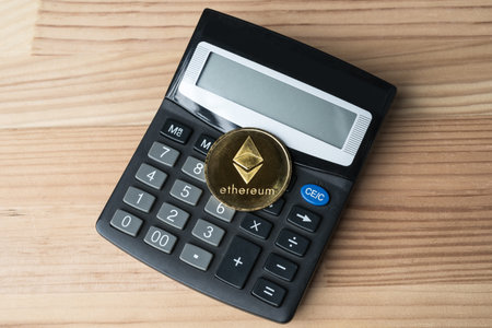 Golden physical Ethereum coin on calculator on wooden background. Cryptocurrency market numbers...