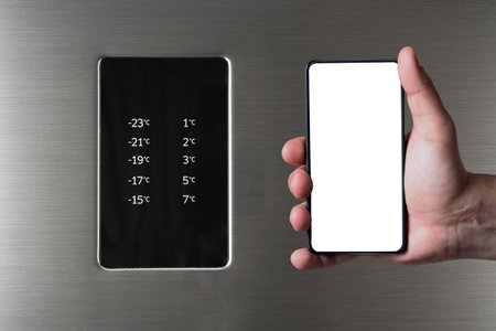 Hand holding frameless smartphone near fridge and freezer control panel. Internet of things abstract...
