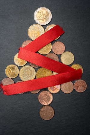 Christmas tree made out of Euro coins with red ribbon chain or garland on dark black slate plate background. Vertical orientation. Flat lay top view.の写真素材