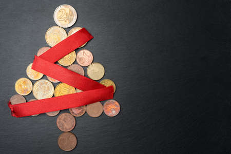Christmas tree made out of Euro coins with red ribbon chain or garland on dark black slate plate background. Copy space on the right. Flat lay top view.の写真素材