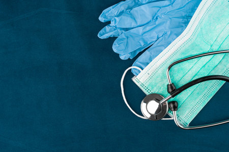 Green protective face mask, stethoscope and gloves on a blue background. Copy space on the left.