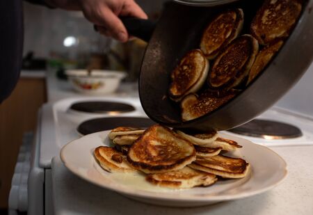 mini pancakes just baked on a cast-iron thick pan will now fall into the plate, where there are already several prepared pancakes. an ordinary kitchen, you can see part of the interior, a hand holding a tilted frying panの写真素材