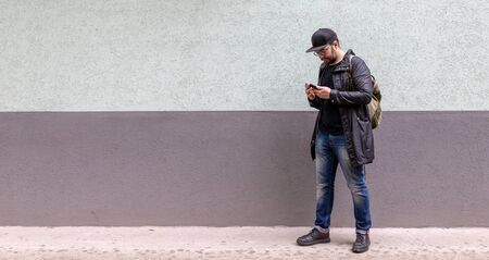 a man with a casual backpack. it stands next to a two-color wall. great background. there is a place for the inscription. the man is handsome. he is dressed in a black raincoat, a black t-shirt, and a cap. the man has a phone in his handの写真素材