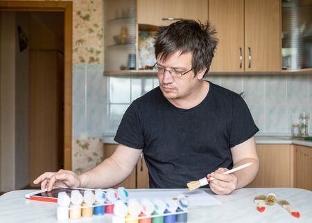 a man at home at the table. in one hand he holds a brush. the other is checked in the tablet. in front of him on the table paints of different colors, brushes.の写真素材