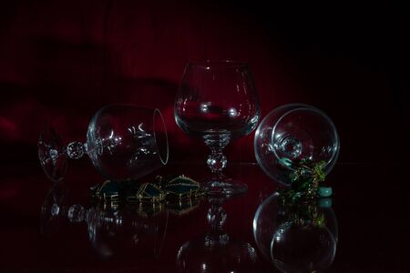 Three necklaces, two of them with stones and one of an unusual alloy that looks like gold. Composition with empty transparent crystal glasses. Everything is reflected in the glass. Dark red backgroundの写真素材