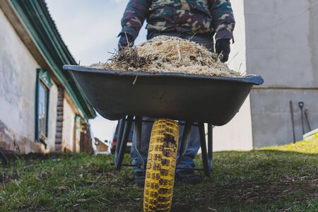 a man in work clothes with a one-wheeled cart in the garden. he rolls it down the slide right at the camera yellow wheel forwardの写真素材