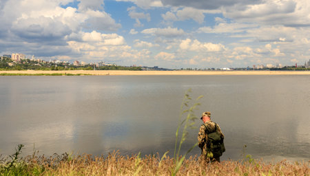 a man dressed in khaki-colored clothes stands on the shore of a reservoir and fishes. buildings and...
