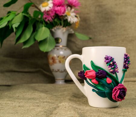 The original mug with decorative figurines made with their own hands. Figures on the cup are fashioned hand made from colored clay. Vase with fresh flowers on a background. High quality photoの写真素材