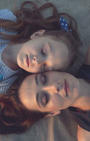 Mom and her daughter are lying on a sand-colored mat, resting. The camera looks at them from above,...