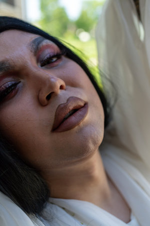 Drag queen posing close to old building in white dress. A wig with Black long hair and make up. High...