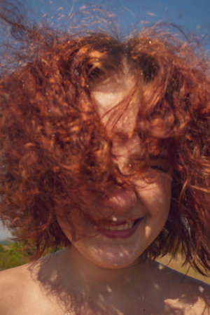 the red-haired curly-haired girl smiles happily and looks at the camera. her thick hair covers her...