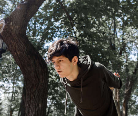 a young guy of Eastern appearance with black hair and a handsome expressive face poses in a Park against the background of trees and architecture. High quality photoの写真素材