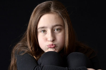 Teenaged girl hugging knees with bored expression wearing black shirt on black background shown from...