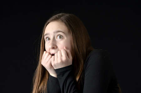 Teenaged girl with hands to mouth and wide eyes in expression of fear wearing black shirt on black backgroundの写真素材