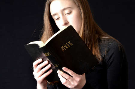 Teenaged girl reading black leather-bound Bible with illuminated face wearing black shirt on black backgroundの写真素材