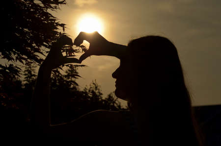 Teen girl in silhouetted profile holding hands in heart shape with sun set and leafy tree backgroundの写真素材