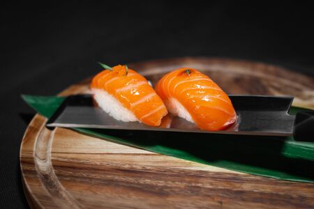 Sushi salmon nigiri with decoration on top, served on japanese knife on round wooden board with dark background.の写真素材